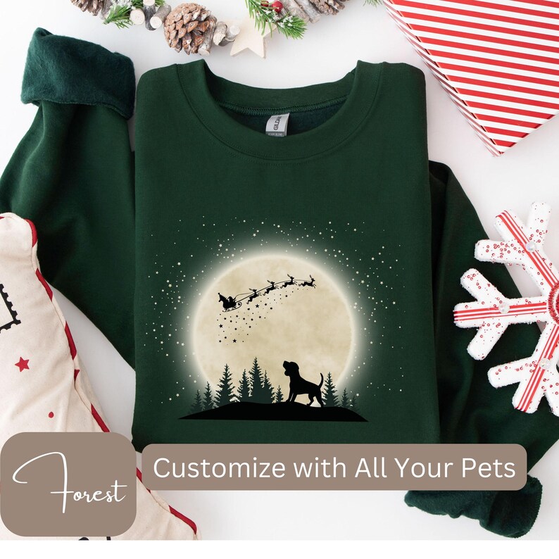 May include: A dark green sweatshirt featuring a silhouette of a dog in front of a full moon with Santa's sleigh and reindeer. The text "Customize with All Your Pets" is visible. The sweatshirt is surrounded by holiday decorations.