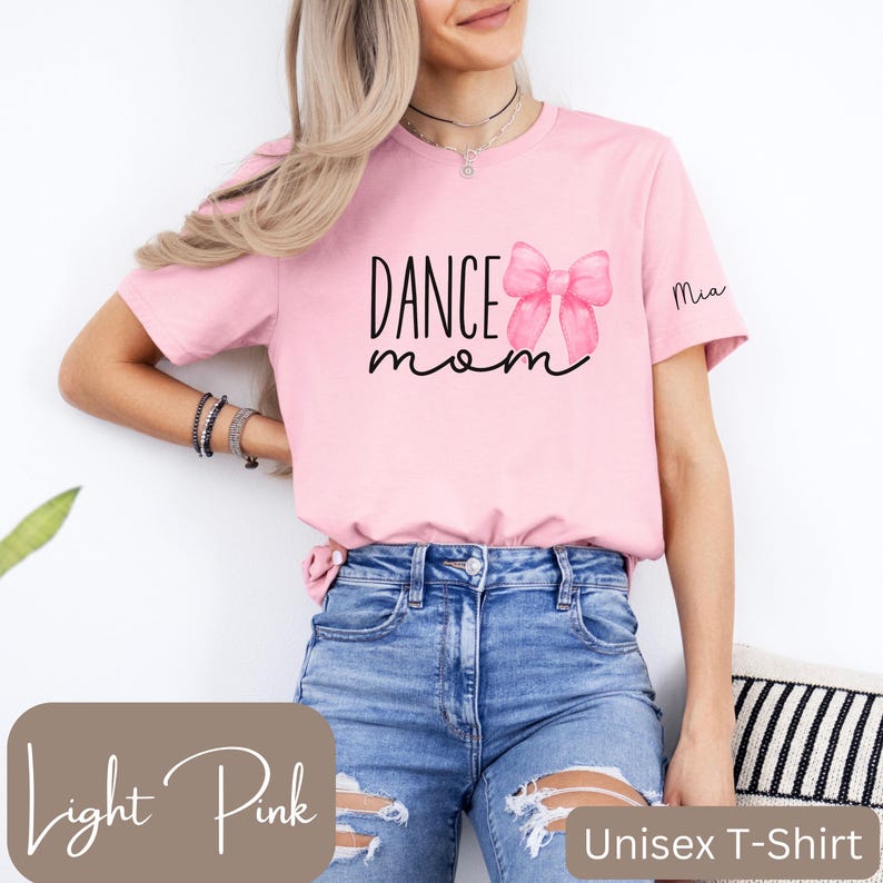 Może przedstawiać: Jasnor&oacute;żowa koszulka unisex z napisem "Dance Mom" i r&oacute;żową kokardką. Na rękawie napisano imię "Mia".