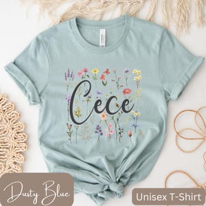 May include: A dusty blue unisex t-shirt with a floral design and the name "Cece" in a black script font.