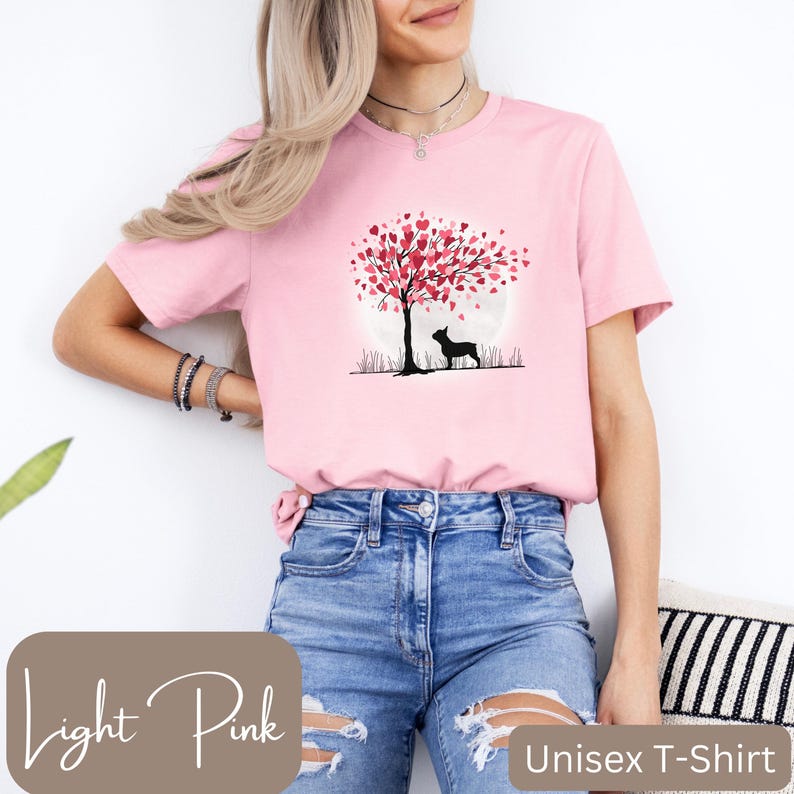 May include: A light pink unisex t-shirt with a graphic of a silhouette of a dog standing under a tree with pink hearts for leaves. The tree is in front of a full moon. The text "Unisex T-Shirt" is at the bottom of the image.