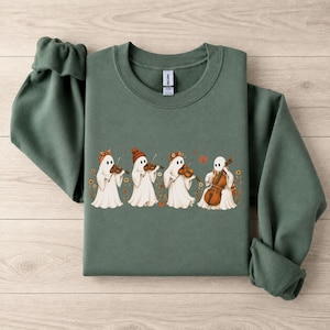 Könnte beinhalten: Ein waldgrüner Rundhals-Pullover mit einer Grafik von vier weißen Geistern, die Streichinstrumente spielen. Die Geister tragen herbstliche Accessoires wie Mützen und Blumenkränze, umgeben von kleinen orangefarbenen Blumen und Blättern.