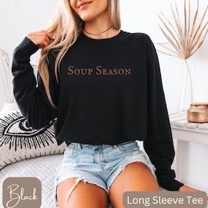 Puede incluir: Camiseta negra de manga larga con el texto "SOUP SEASON" en fuente marr&oacute;n. La camiseta se combina con pantalones cortos de mezclilla. Las palabras "Black" y "Long Sleeve Tee" son visibles en la imagen.