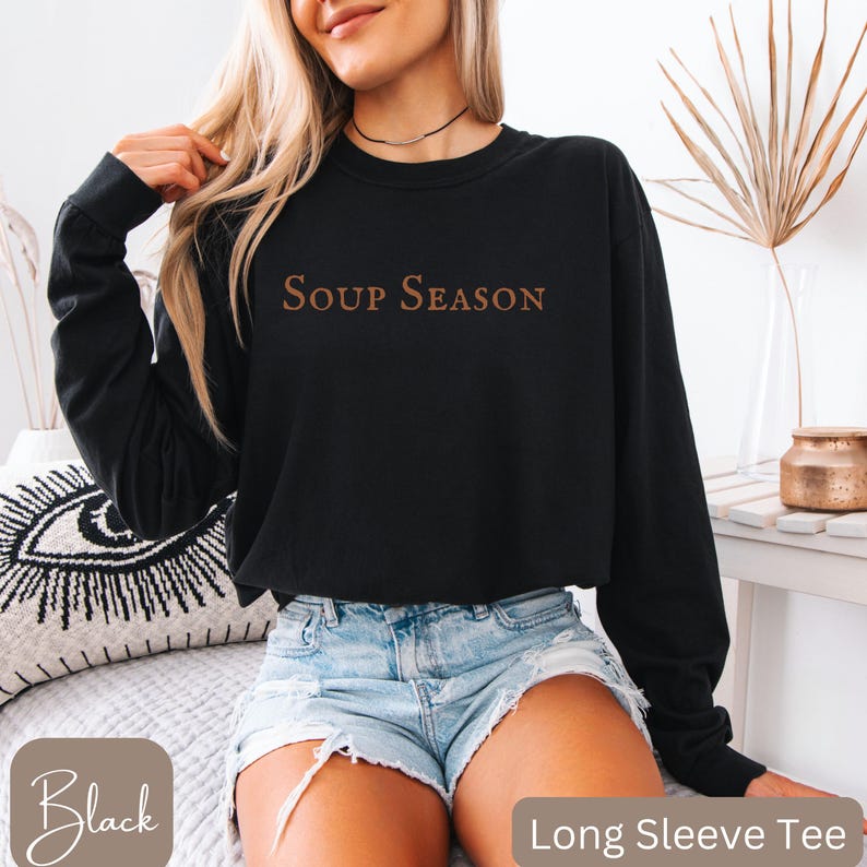 Puede incluir: Camiseta negra de manga larga con el texto "SOUP SEASON" en fuente marr&oacute;n. La camiseta se combina con pantalones cortos de mezclilla. Las palabras "Black" y "Long Sleeve Tee" son visibles en la imagen.