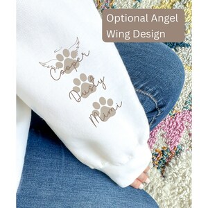 May include: White sweatshirt with brown paw prints and names "Cooper", "Dusty", and "Mimi" printed on the sleeve. The text "Optional Angel Wing Design" is printed on the top of the sleeve.