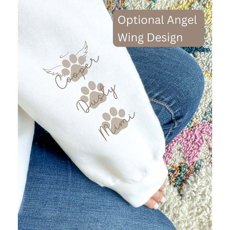 May include: White sweatshirt with brown paw prints and names "Cooper", "Dusty", and "Mimi" printed on the sleeve. The text "Optional Angel Wing Design" is printed on the top of the sleeve.