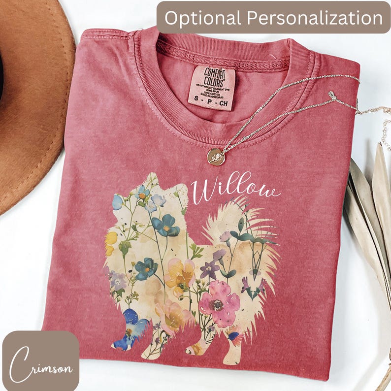 May include: Crimson-colored t-shirt with a floral dog design and the name "Willow" in white script. The shirt has a crew neck and short sleeves. A silver necklace with a pendant is draped across the shirt. The words "Optional Personalization" are at the top.