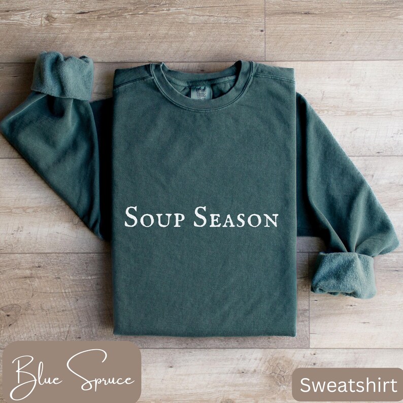 Puede incluir: Una sudadera de color verde azulado con la frase "Soup Season" en blanco. Las mangas est&aacute;n dobladas. Tambi&eacute;n se ven las palabras "Blue Spruce" y "Sweatshirt". La prenda est&aacute; sobre una superficie de madera.