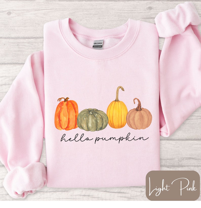 May include: Light pink sweatshirt with a watercolor design of four pumpkins in orange, green, yellow, and brown. The words "hello pumpkin" are written in a black script font below the pumpkins. The sweatshirt is a Gildan brand.