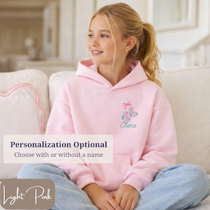 May include: Light pink hoodie with a drawstring hood. The front features an embroidered design of ice skates with a bow and the name "Clara" in teal. The text "Personalization Optional" and "Light Pink" are also visible.