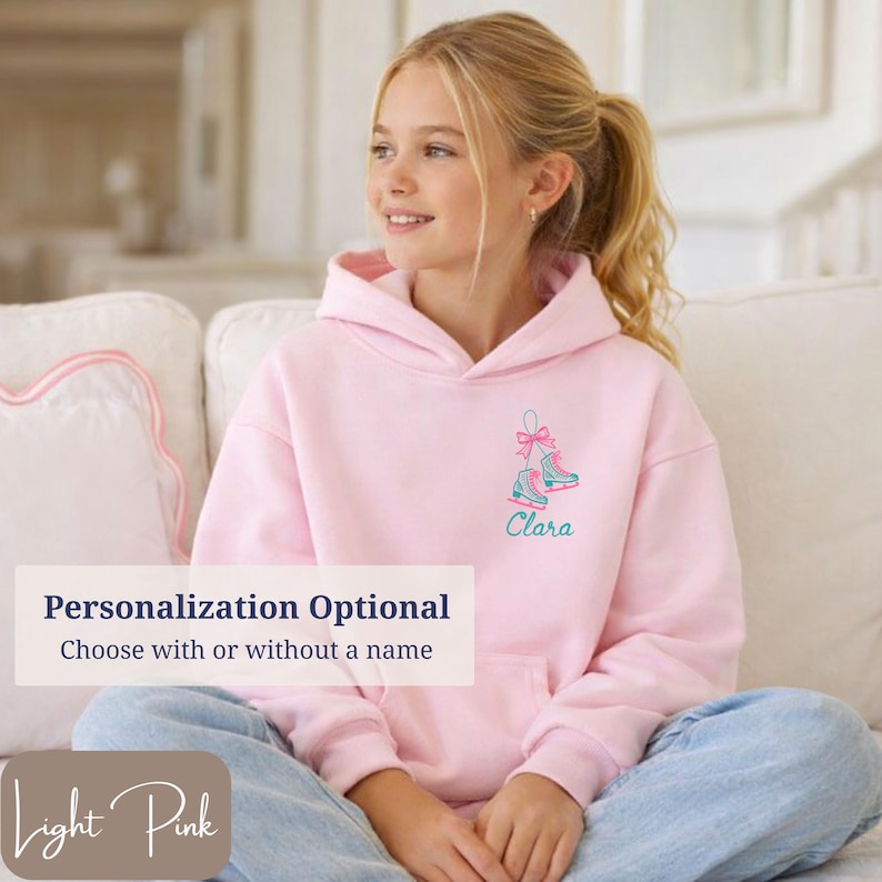 May include: Light pink hoodie with a drawstring hood. The front features an embroidered design of ice skates with a bow and the name "Clara" in teal. The text "Personalization Optional" and "Light Pink" are also visible.
