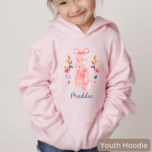 May include: A light pink youth hoodie featuring a watercolor design of ballet shoes with a bow and floral accents. The name "Maddie" is written in a cursive font below the ballet shoes. The hoodie has a front pocket and a hood.