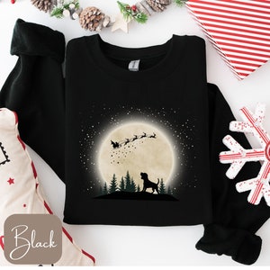 May include: Black sweatshirt featuring a silhouette of a dog looking up at Santa Claus in his sleigh, flying across a full moon. The design includes a forest of trees and a starry sky. The word "Black" is in a box in the lower left corner.