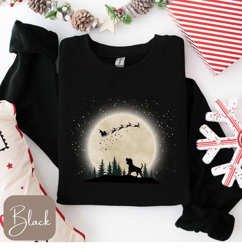May include: Black sweatshirt featuring a silhouette of a dog looking up at Santa Claus in his sleigh, flying across a full moon. The design includes a forest of trees and a starry sky. The word "Black" is in a box in the lower left corner.