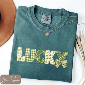 May include: Teal Comfort Colors t-shirt with the word "LUCKY" in a patchwork design. The letters feature green gingham, floral, and polka dot patterns. A gold necklace with a pendant rests on the shirt. The shirt has a small tag that says "COMFORT COLORS".