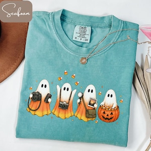 May include: A seafoam green short-sleeve t-shirt featuring a graphic of four ghosts with candy corn bodies. The ghosts are holding cameras, a photograph, and a jack-o'-lantern filled with candy corn. Small candy corn shapes surround them.
