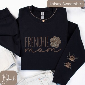 May include: Black unisex sweatshirt with the text "Frenchie mom" in brown lettering. A paw print with a leopard print design is above the text. The right sleeve has the text "Cooper" and "Dixie" with paw prints and a wing.