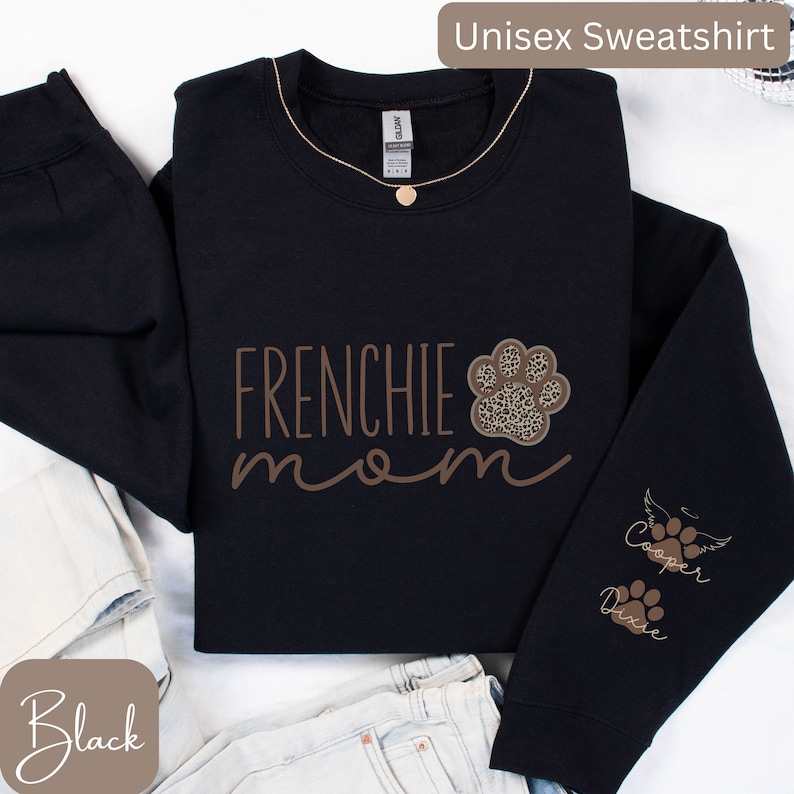 May include: Black unisex sweatshirt with the text "Frenchie mom" in brown lettering. A paw print with a leopard print design is above the text. The right sleeve has the text "Cooper" and "Dixie" with paw prints and a wing.