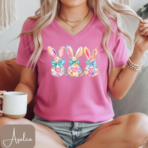Op de afbeelding: Een roze V-hals T-shirt met een aquarelontwerp van drie paashazen. Elke konijn heeft een bloemenpatroon, een hart en een blauwe strik. Het woord "Azalea" staat in de linkerbenedenhoek.