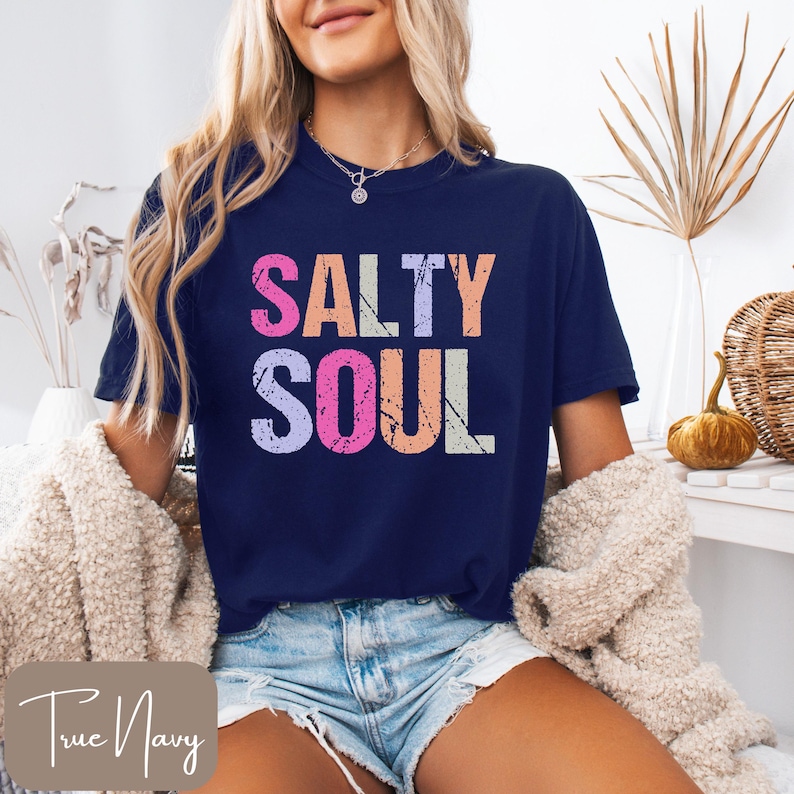 May include: Navy blue t-shirt with the words "SALTY SOUL" in colorful, distressed lettering. The shirt is a crew neck style and is paired with denim shorts. A cream-colored sweater is draped over the shoulders.