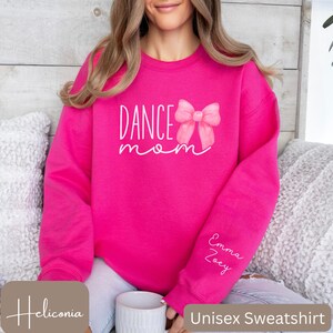 Może przedstawiać: R&oacute;żowa bluza unisex z napisem "Dance Mom" i r&oacute;żową wstążką w stylu akwareli. Bluza ma napisane na rękawie imiona "Emma" i "Zoey".