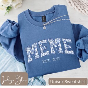 Op de afbeelding: Indigo blauwe unisex sweatshirt met het woord "MEME" in witte bloemenprint letters. De tekst "EST. 2025" staat onder het woord "MEME". Een zilveren ketting met een hanger ligt op de sweatshirt. De sweatshirt is op een denim oppervlak gevouwen.