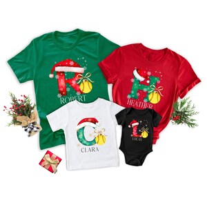 Op de afbeelding: Vier kerst-t-shirts en een romper. De shirts zijn groen, rood, wit en zwart. Elk shirt heeft een letter met een kerstmuts, sneeuwvlokken en een honkbal. De namen "Robert", "Heather", "Clara" en "Lucas" staan op de shirts.