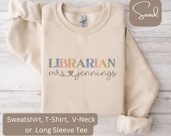 May include: A beige sweatshirt with the text "LIBRARIAN mrs ♥ jennings" in colorful letters. The sweatshirt is available in a variety of styles, including a sweatshirt, t-shirt, v-neck, and long sleeve tee.