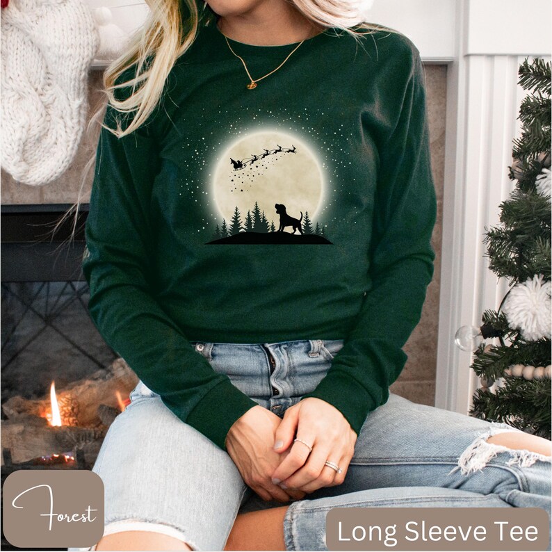 May include: A dark green long-sleeve tee with a graphic of a dog silhouette under a full moon with Santa's sleigh and reindeer. The shirt has the text "Long Sleeve Tee" at the bottom.