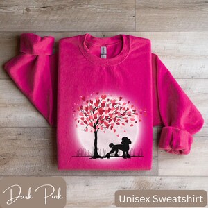 Op de afbeelding: Een donkerroze unisex sweatshirt met een afbeelding van een poedel die onder een boom staat met harten als bladeren. De boom staat voor een volle maan.