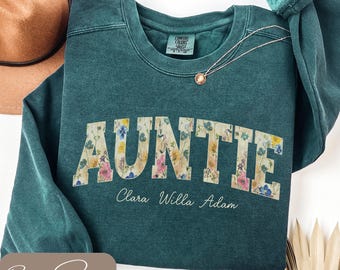 May include: A teal sweatshirt featuring the word "AUNTIE" in large, floral-filled letters. Below the word are the names "Clara Willa Adam" in a script font. A gold necklace with a pendant is visible.