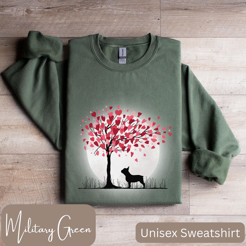 May include: A unisex sweatshirt in military green featuring a silhouette of a French Bulldog standing in front of a tree with pink hearts for leaves. The tree is in front of a white moon.