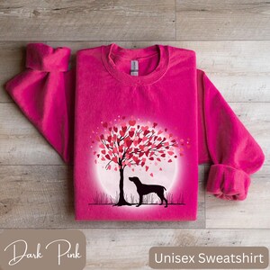 May include: A dark pink unisex sweatshirt featuring a graphic of a dog standing beneath a tree with hearts for leaves. The tree is encircled by a white circle with a light pink gradient.