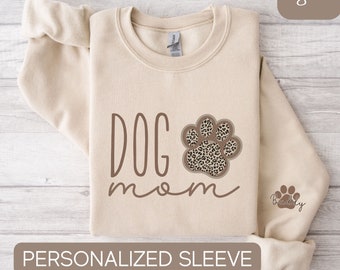 May include: A beige sweatshirt with the text "DOG mom" in brown lettering and a brown paw print with a leopard print design. The sweatshirt has a personalized sleeve with a small paw print and the word "Buddy" in brown lettering.
