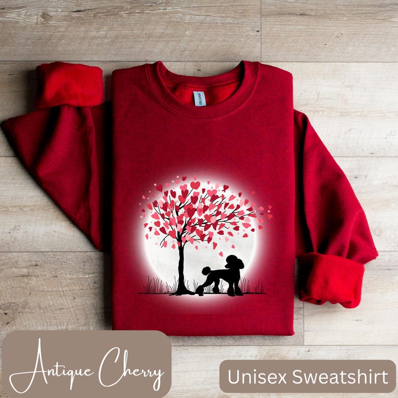 Op de afbeelding: Rood unisex sweatshirt met een afbeelding van een poedel die onder een boom staat met hartvormige bladeren. De boom staat voor een witte maan. De tekst "Antique Cherry" staat linksonder en "Unisex Sweatshirt" rechtsonder.