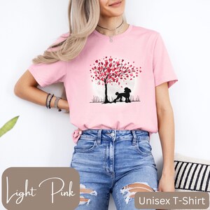 Op de afbeelding: Een lichtroze unisex t-shirt met een afbeelding van een poedel die onder een boom staat met roze hartvormige bladeren. De tekst "Light Pink" is zichtbaar in de linkerbenedenhoek van de afbeelding. De tekst "Unisex T-Shirt" is zichtbaar in de rechterbenedenhoek van de afbeelding.