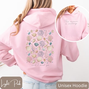 K&ouml;nnte beinhalten: Hellrosa unisex Hoodie mit einem Muschel- und Korallendesign auf der R&uuml;ckseite. Der Text "The ocean is calling and I must go" ist auf der R&uuml;ckseite des Hoodies aufgedruckt.