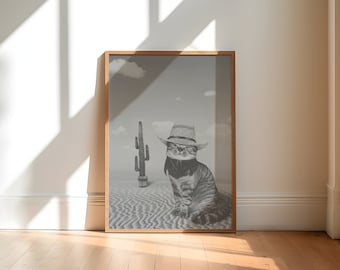 May include: A black and white photograph of a cat wearing a cowboy hat and sunglasses, sitting in a desert landscape with a cactus in the background.