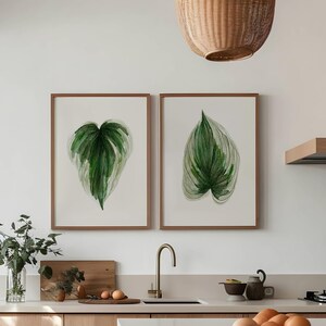 May include: A modern kitchen with light wood cabinets and a white countertop. Two framed watercolor paintings of green leaves hang on the wall above the countertop. A wicker chair sits in front of the island.