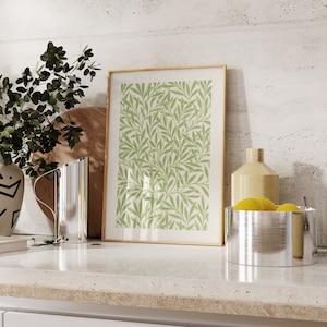 May include: A framed print with a green leaf pattern on a white background. The print is sitting on a worktop with a silver jug, a wooden chopping board, a yellow vase, and a silver bowl with lemons.