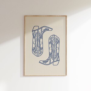 Op de afbeelding: Een printbare kunstprint met een lijntekening van twee cowboylaarzen met ingewikkelde ontwerpen. De laarzen zijn omlijnd in marineblauw tegen een beige achtergrond.