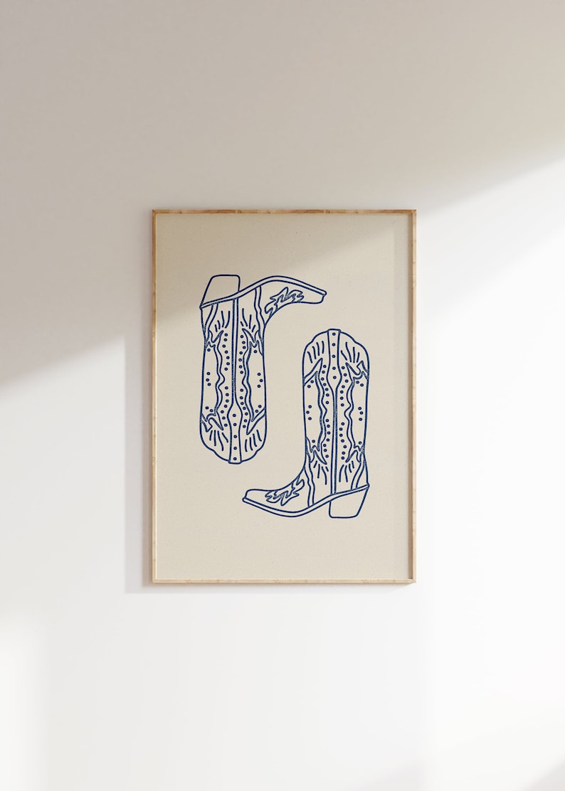 Op de afbeelding: Een printbare kunstprint met een lijntekening van twee cowboylaarzen met ingewikkelde ontwerpen. De laarzen zijn omlijnd in marineblauw tegen een beige achtergrond.