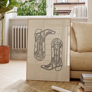 May include: A framed black and white line drawing of two cowboy boots with intricate designs. The boots are on a beige background.