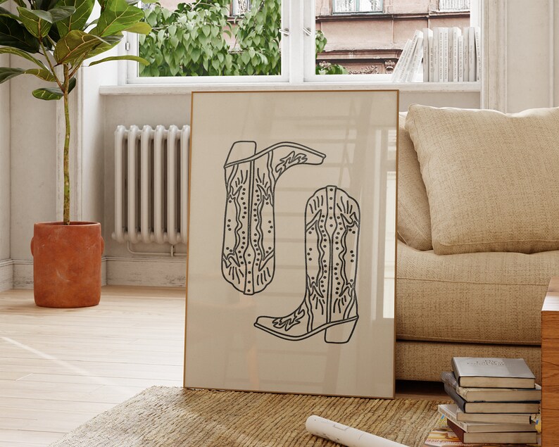 May include: A framed black and white line drawing of two cowboy boots with intricate designs. The boots are on a beige background.