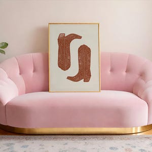 Pu&ograve; includere: Un divano in velluto rosa con un bordo dorato si trova di fronte a un muro bianco. Una stampa incorniciata di due stivali da cowboy marroni con un motivo geometrico &egrave; appesa sopra il divano.