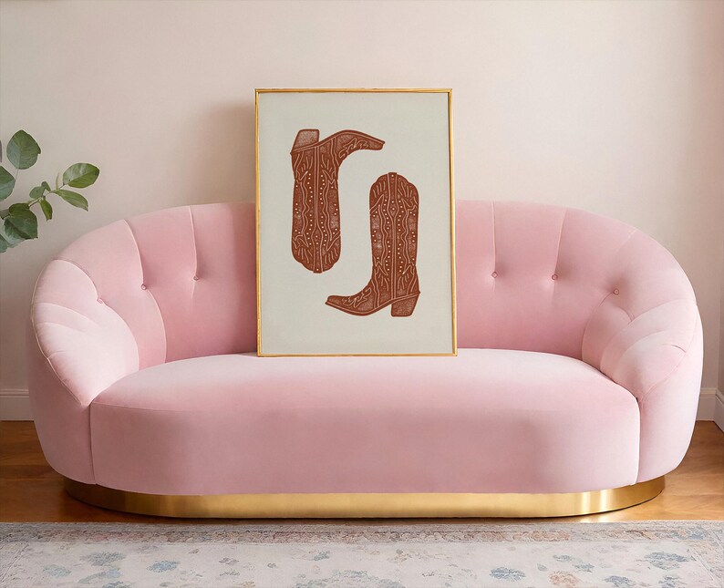 Pu&ograve; includere: Un divano in velluto rosa con un bordo dorato si trova di fronte a un muro bianco. Una stampa incorniciata di due stivali da cowboy marroni con un motivo geometrico &egrave; appesa sopra il divano.