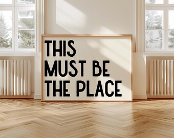 May include: A framed art print with the text "THIS MUST BE THE PLACE" in bold black letters on a cream background. The artwork is displayed on a wooden floor, with windows and radiators in the background.