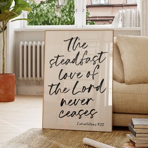 May include: A beige framed print with black calligraphy that reads "The steadfast love of the Lord never ceases, Lamentations 3:22".