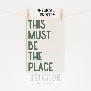May include: A cream-colored print with the text "THIS MUST BE THE PLACE" in dark green, held by wooden clothespins on a string. The words "PHYSICAL PRINT!" are above the text, and "BUNGALOW print shop" is below.