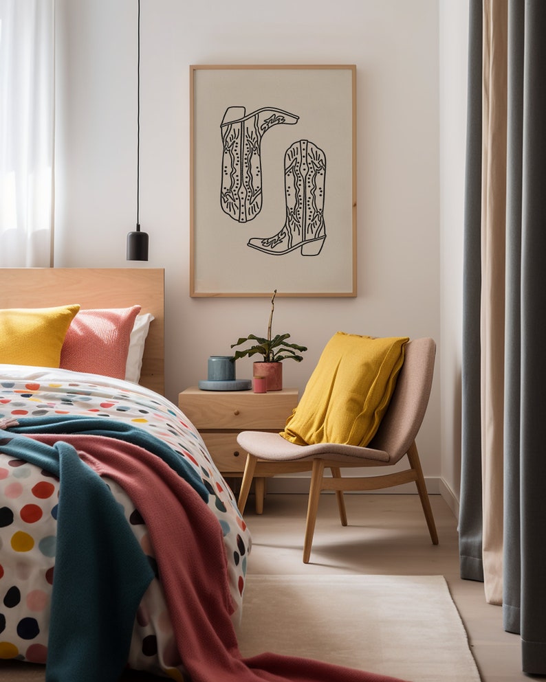 May include: A bedroom with a bed, a chair, a nightstand, and a framed print of two cowboy boots. The bed has a white and colorful polka dot duvet cover, a pink blanket, and a blue blanket. The chair is wooden with a yellow pillow. The nightstand is wooden with a small plant on top. The print is on a beige background with black outlines.