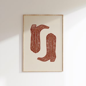 Pu&ograve; includere: Una stampa incorniciata di due stivali da cowboy marroni con disegni intricati. Gli stivali sono su uno sfondo crema.